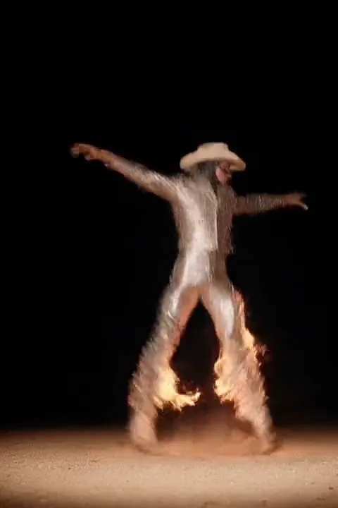 Dancer in a fringed white outfit and cowboy hat spinning through flames and dust at night