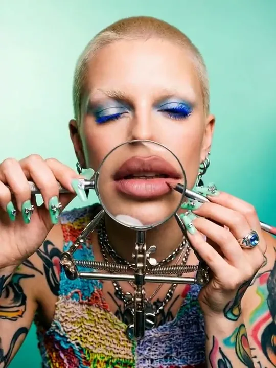Buzzcut model with blue mascara holding a magnifying glass over her lips, mint backdrop