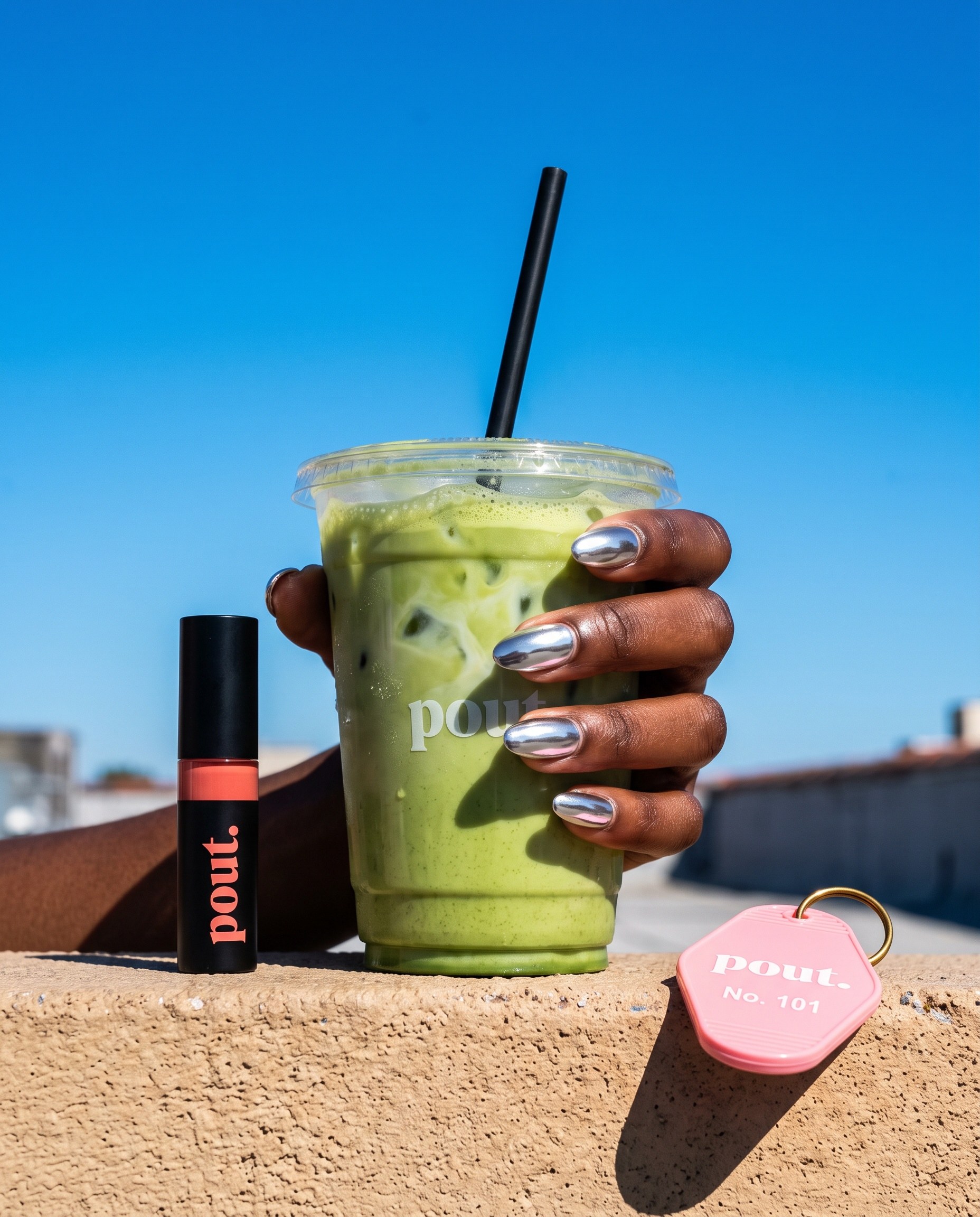 Chrome-nailed hand holding an iced matcha with pout. lip tint and motel keychain against a blue sky