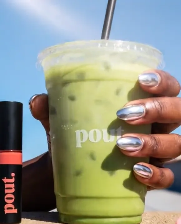 Chrome-nailed hand holding an iced matcha with pout. lip tint and motel keychain against a blue sky