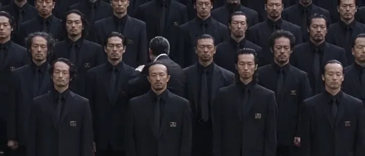 Ultra-wide shot of a crowd of men in black suits, one turning against the flow