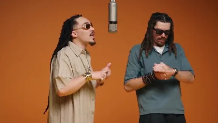 Two men with dreadlocks performing into a hanging studio microphone against an orange backdrop