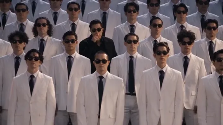 Man in a black suit standing among a crowd of identical men in white suits and sunglasses